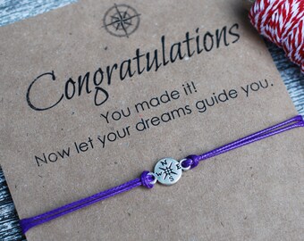 May include: A purple friendship bracelet with a silver compass charm and a card that reads "Congratulations You made it! Now let your dreams guide you."
