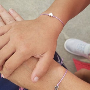 May include: Two people wearing matching friendship bracelets with small silver heart charms on a light purple cord.