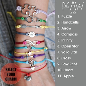 May include: A close-up of a person's wrist wearing eleven colorful bracelets with different charms. The charms include a puzzle, handcuffs, arrow, compass, infinity symbol, open star, solid star, cross, paw print, heart, and apple. The bracelets are made of string and the charms are silver.