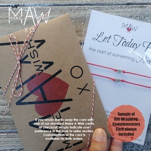 Puede incluir: Una tarjeta de papel kraft con el texto "Make A Wish" y un coraz&oacute;n rojo. La tarjeta est&aacute; atada con un cordel rojo y blanco. Una tarjeta separada con el texto "Let Today Be the start of something New" y dos pulseras de cuerda roja con dijes de coraz&oacute;n plateados. El texto "Sample of Gift Wrapping. Complementary Card always Included" es visible.