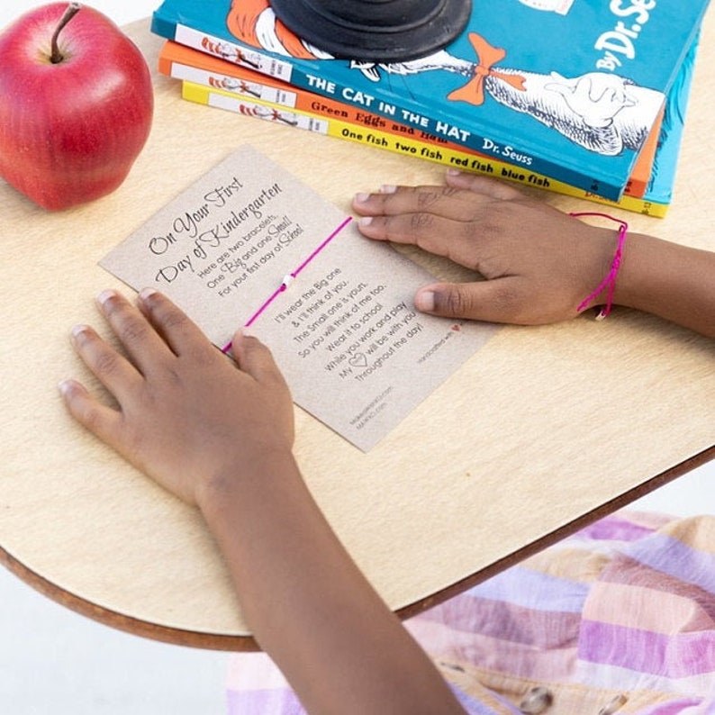 May include: A pink and white friendship bracelet with a card that reads "On Your First Day of Kindergarten" and a message about wearing the bracelets to school. The card is on a wooden table with a red apple and books in the background.