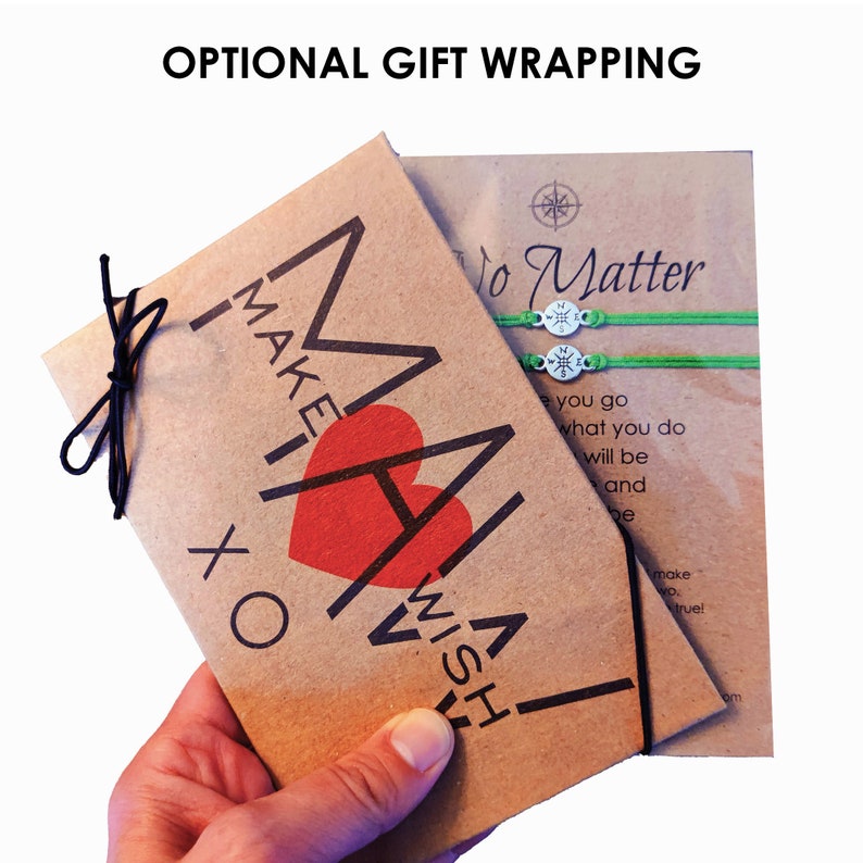 Pu&ograve; includere: Un sacchetto regalo di carta marrone con un cuore rosso e il testo "MAKE A WISH" stampato su di esso. Il sacchetto &egrave; legato con uno spago nero ed &egrave; tenuto da una mano. Il sacchetto &egrave; accanto a un biglietto con il testo "No Matter" e due braccialetti verdi con ciondoli a forma di bussola.