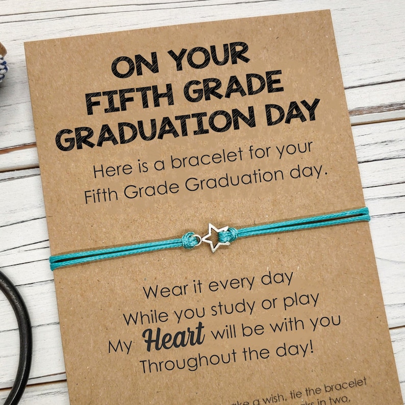 Puede incluir: Una tarjeta marr&oacute;n con el texto "On Your Fifth Grade Graduation Day" y un brazalete de cuerda turquesa con un dije de estrella plateada. La tarjeta dice "Here is a bracelet for your Fifth Grade Graduation day. Wear it every day while you study or play. My Heart will be with you Throughout the day!"