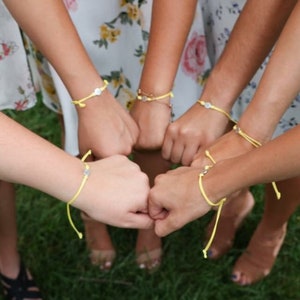 May include: A group of friends wearing yellow friendship bracelets with silver charms. The bracelets are tied around their wrists and they are holding their hands together in a circle.