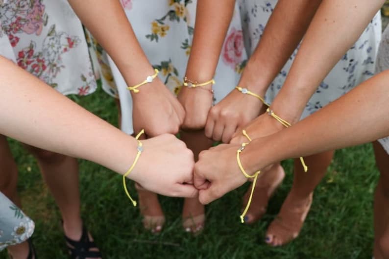 May include: A group of friends wearing yellow friendship bracelets with silver charms. The bracelets are tied around their wrists and they are holding their hands together in a circle.