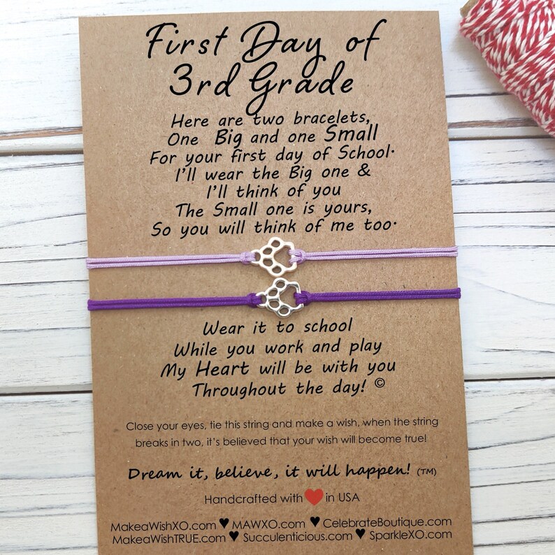 May include: A card with a message for a child starting 3rd grade. The card includes two purple and lavender friendship bracelets with a silver paw print charm. The card reads "First Day of 3rd Grade" and includes a message about the bracelets and a wish-making tradition.