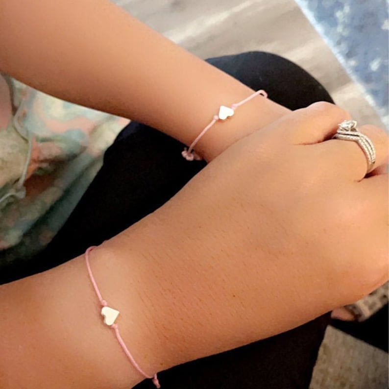 May include: Two pink friendship bracelets with silver heart charms. The bracelets are tied on the wrists of two people.