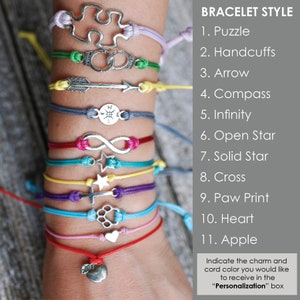 Pu&ograve; includere: Primo piano di un polso con undici braccialetti colorati con diversi ciondoli d'argento. I ciondoli includono un puzzle, manette, una freccia, una bussola, un simbolo di infinito, una stella aperta, una stella piena, una croce, un'impronta di zampa, un cuore e una mela. I braccialetti sono realizzati in cordoncino e sono disponibili in una variet&agrave; di colori.