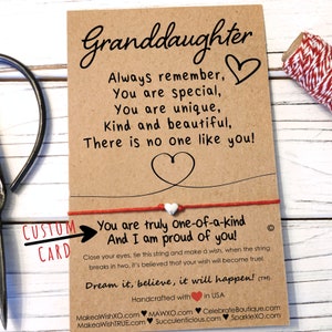May include: A brown card with a red string tied around it. The card says "Granddaughter" and has a message about being special and unique. The string has a small white bead on it. The card also says "You are truly one-of-a-kind And I am proud of you!" and "Dream it, believe, it will happen!"