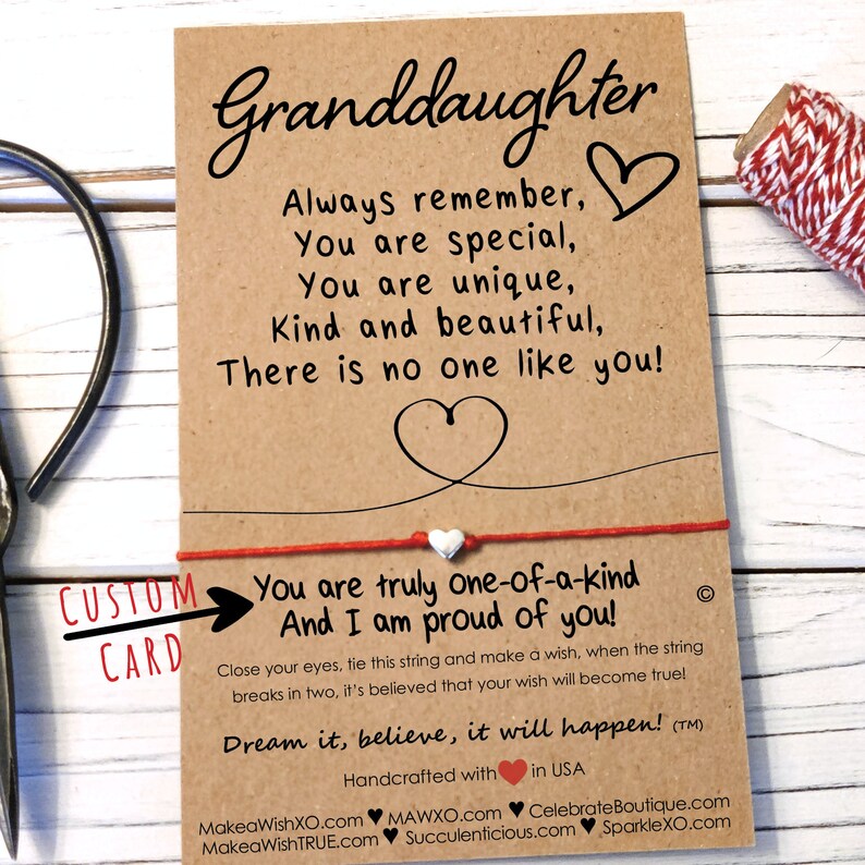 May include: A brown card with a red string tied around it. The card says "Granddaughter" and has a message about being special and unique. The string has a small white bead on it. The card also says "You are truly one-of-a-kind And I am proud of you!" and "Dream it, believe, it will happen!"
