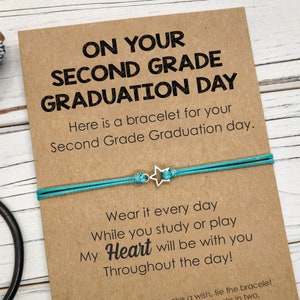 Puede incluir: Una tarjeta con el texto "On Your Second Grade Graduation Day" y una pulsera de amistad turquesa con un dije de estrella plateada. La tarjeta dice "Here is a bracelet for your Second Grade Graduation day. Wear it every day while you study or play. My Heart will be with you throughout the day!"