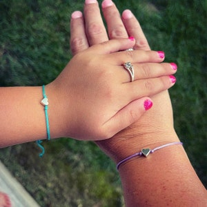 May include: Two hands, one with a silver ring and the other with a turquoise bracelet with a silver heart charm, are holding each other. The hand with the ring also has a purple bracelet with a silver heart charm.