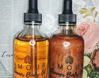 May include: Two amber glass bottles with dropper tops filled with shimmer body oil. The bottles have black labels with white text that reads "Amour Shimmer Body Oil" and "Botanicals Beauty". The labels also state "No Parabens, Phthalates, Silicones, Sulfates, Petroleum Chemicals".