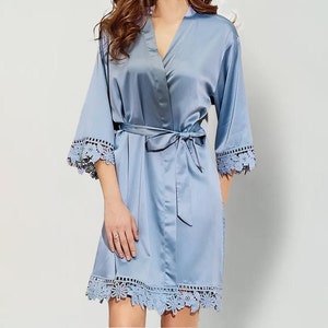 May include: A light blue satin robe featuring a V-neckline, three-quarter length sleeves, and a matching tie belt at the waist. The sleeve cuffs and the bottom hem are finished with a decorative floral lace trim in a matching light blue colour.