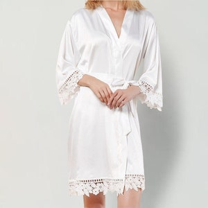 May include: A white satin robe featuring a V-neckline, long sleeves, and a matching tie belt at the waist. The sleeve cuffs and the bottom hem are decorated with intricate white floral lace trim, providing a detailed finish to the garment.