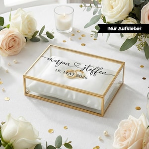 Peut inclure: Boîte à alliances rectangulaire en verre avec un cadre doré. À l'intérieur, deux alliances dorées reposent sur un coussin en satin blanc. La boîte est personnalisée avec les noms "marjan & steffen" et la date "19. MAI 2026".