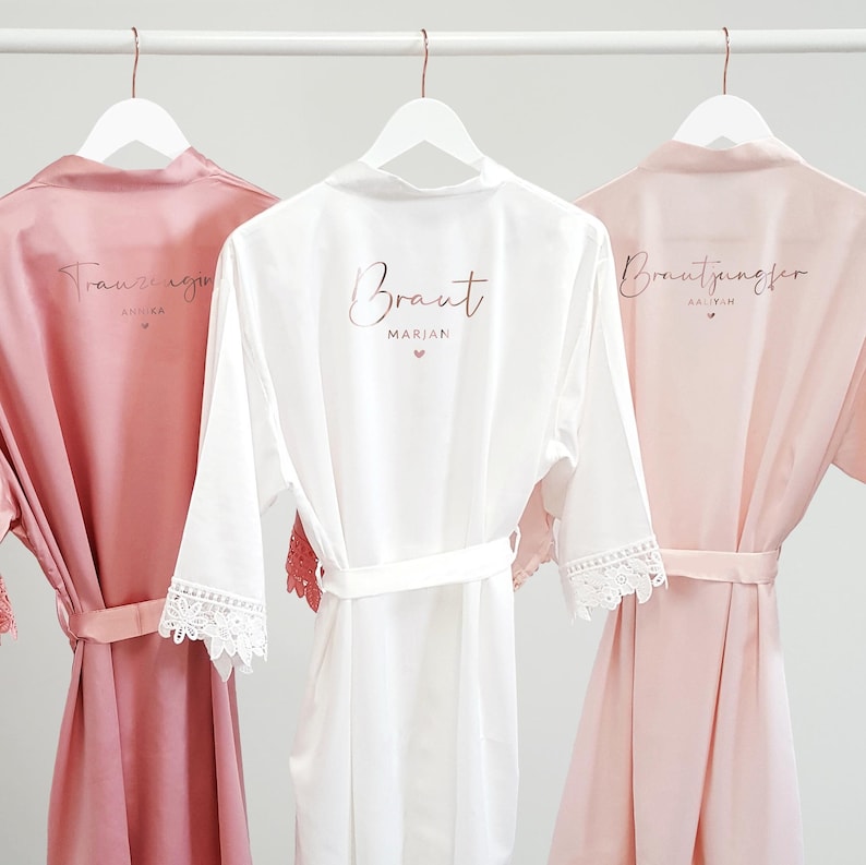 May include: Three satin robes in dusty rose, white, and light pink, each featuring a tie waist and lace-trimmed sleeves. The robes are printed with rose gold text including the words Trauzeugin, Braut, and Brautjungfer, along with names.
