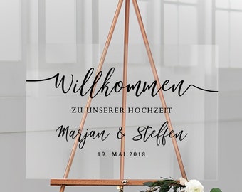 Op de afbeelding: Een helder acryl huwelijkswelkombord met zwarte kalligrafietekst die "Willkommen zu unserer Hochzeit Marjan & Steffen 19. Mai 2018" luidt. Het bord staat op een houten ezel.