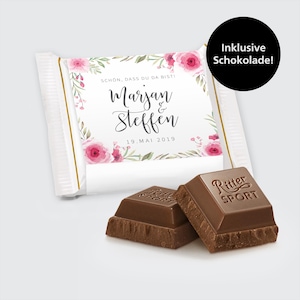 Könnte beinhalten: Weisse Schokoladentafelverpackung mit rosafarbenem Blumendesign und dem Text "SCHON, DASS DU DA BISTI Marjan & Steffen 19. MAI 2019". Die Verpackung liegt neben zwei Ritter Sport Schokoladentafeln.