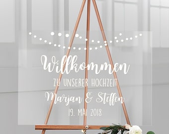 Op de afbeelding: Een helder acryl huwelijksbord met witte letters met de tekst "Willkommen zu unserer Hochzeit Marjan & Steffen 19. Mai 2018". Het bord staat op een houten ezel.