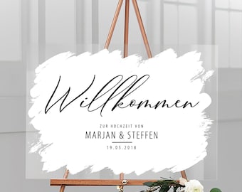 Op de afbeelding: Een wit acryl welkomsbord met zwarte kalligrafie waarop "Willkommen" en "Zur Hochzeit von Marjan & Steffen 19.05.2018" staat. Het bord staat op een houten ezel.