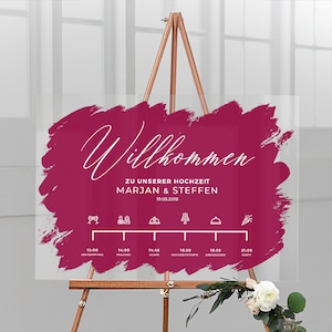 Peut inclure: Panneau de mariage transparent sur chevalet en bois. Le panneau présente le mot "Willkommen" en écriture blanche sur un fond magenta. Les noms Marjan & Steffen et la date sont affichés. Une chronologie avec des icônes d'événements est également présente.