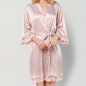 May include: A light pink satin robe featuring long sleeves with wide lace trim at the cuffs and a matching lace border along the hem. The garment includes a wrap-front design with a coordinating fabric tie belt at the waist to secure the closure.