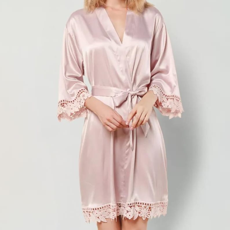 May include: A light pink satin robe featuring long sleeves with wide lace trim at the cuffs and a matching lace border along the hem. The garment includes a wrap-front design with a coordinating fabric tie belt at the waist to secure the closure.