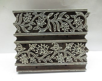 Pode incluir: Um bloco de madeira castanha escura com um desenho floral esculpido. O desenho apresenta flores e folhas brancas. O bloco retangular tem uma borda decorativa, ideal para impressão têxtil ou projetos de artesanato.