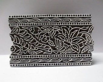 Pode incluir: Um bloco de impressão de madeira preto com um desenho floral branco. O desenho apresenta folhas e flores intrincadas com um padrão de borda repetitivo.