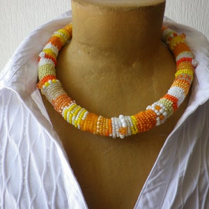 Könnte beinhalten: Eine dicke, mehrfarbige Perlenkette mit orangefarbenen, gelben und weißen Perlen. Die Kette wird auf einem weißen Hemd präsentiert.
