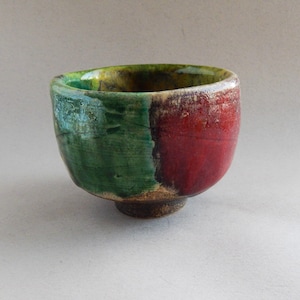 May include: A small, round ceramic bowl with a green, red, and brown glaze. The bowl has a slightly uneven rim and a rounded base.