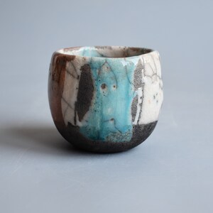 May include: A small, round ceramic cup with a blue, white, and brown glaze. The cup has a rustic, handmade look.