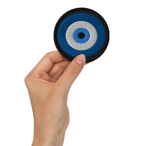 Könnte beinhalten: Ein blauer und weißer böser Blick-Anhänger mit schwarzem Umriss, auf einen schwarzen Stoffkreis genäht.
