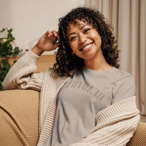 May include: A woman wearing a gray t-shirt with the word "Breathe" printed on it in a gray color. She is wearing a cream cardigan and has curly brown hair.