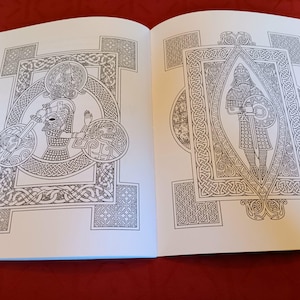 Puede incluir: Libro abierto con dos ilustraciones detalladas en blanco y negro. Las ilustraciones presentan intrincados dise&ntilde;os de nudos celtas, enmarcando figuras con escudos, espadas y cascos. Las p&aacute;ginas son blancas, sobre un fondo rojo oscuro.