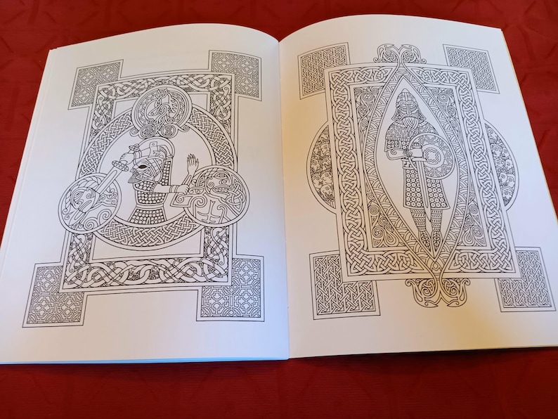 Puede incluir: Libro abierto con dos ilustraciones detalladas en blanco y negro. Las ilustraciones presentan intrincados dise&ntilde;os de nudos celtas, enmarcando figuras con escudos, espadas y cascos. Las p&aacute;ginas son blancas, sobre un fondo rojo oscuro.