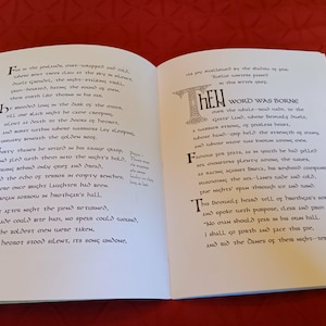 Puede incluir: Libro abierto con texto impreso en una fuente estilizada. Las letras iniciales son decorativas. El libro est&aacute; sobre una superficie roja. El texto parece ser un poema o una obra literaria.