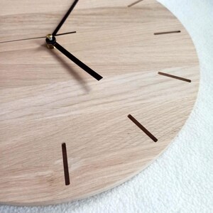 May include: A close-up of a round wooden clock with a light wood grain finish. The clock has black hands and simple brown line markers for the hours. The clock's hands are positioned to show the time.