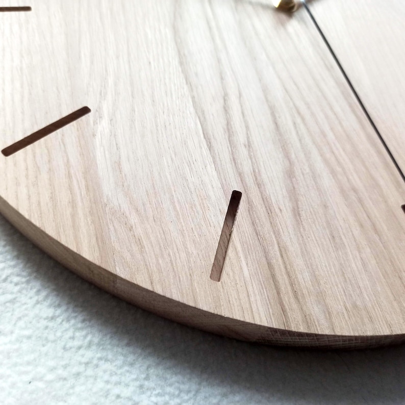 May include: Close-up of a wooden clock face. The clock is made of light-colored wood with a natural grain. The clock face has simple, minimalist markings, with thin, dark lines indicating the hours. The hands are thin and black.
