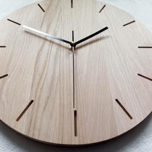 May include: A round wooden clock with a light brown finish. The clock face has minimalist black hands and hour markers. The clock's hands are black and silver, and the hour markers are thin, cut-out lines.