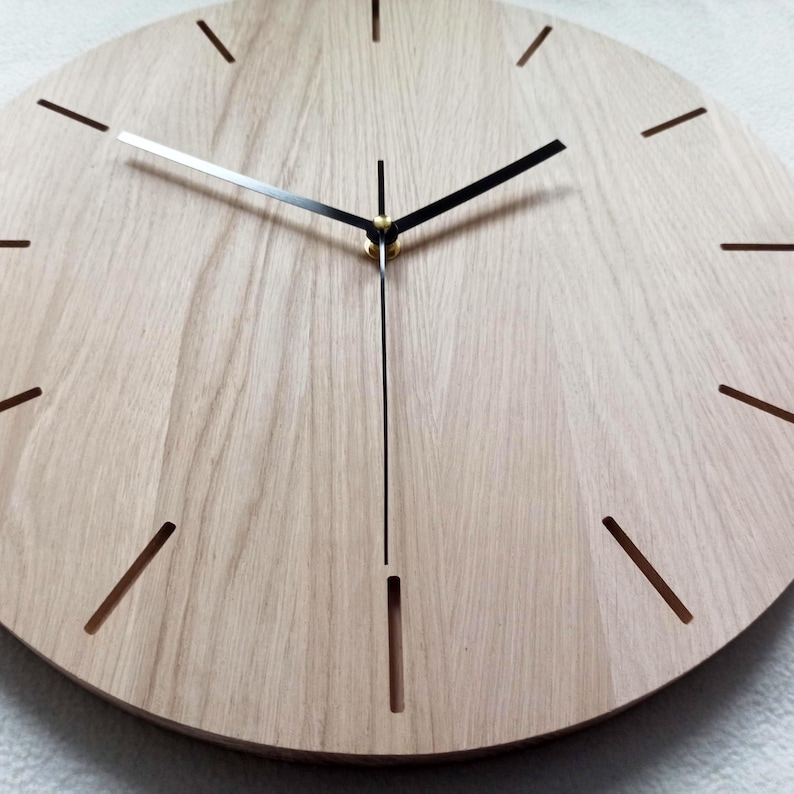 May include: A round wooden clock with a light brown finish. The clock face has minimalist black hands and hour markers. The clock's hands are black and silver, and the hour markers are thin, cut-out lines.