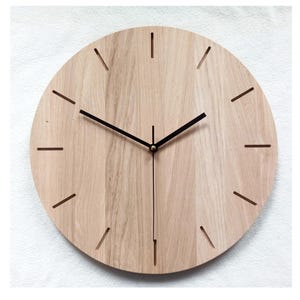 May include: A round wooden wall clock with a light brown finish. The clock face features black hands and simple line markers for the hours. The clock has a minimalist design, suitable for various interior styles.