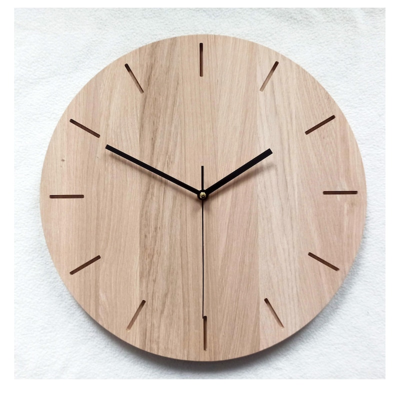 May include: A round wooden wall clock with a light brown finish. The clock face features black hands and simple line markers for the hours. The clock has a minimalist design, suitable for various interior styles.