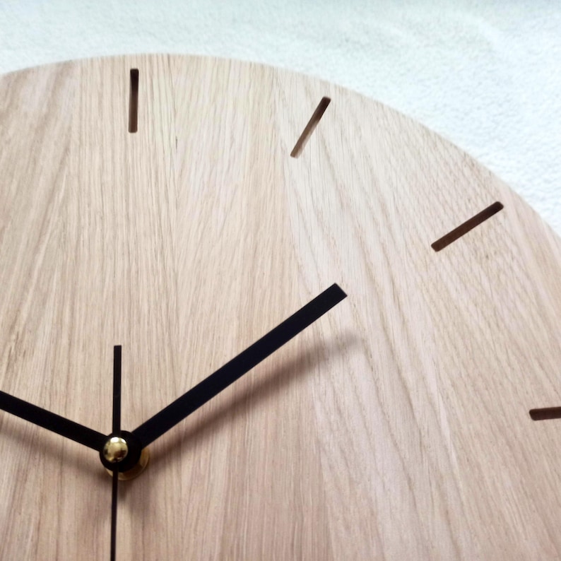 May include: Close-up of a light-colored wooden clock face with black hands and hour markers. The clock has a minimalist design, with a natural wood grain finish. The hands are black and the hour markers are simple lines.