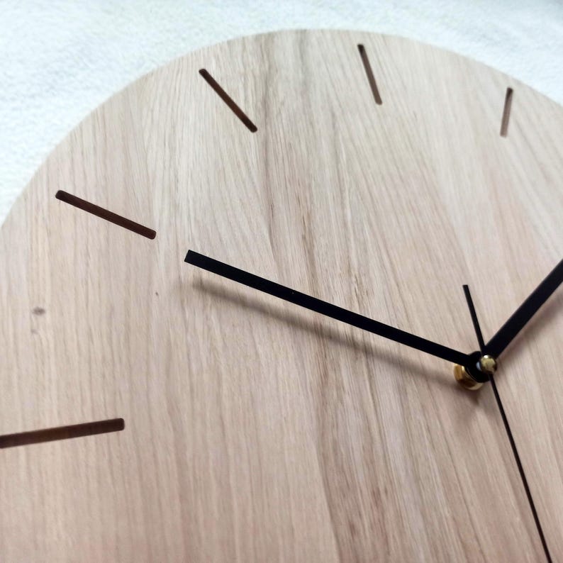 May include: Close-up of a wooden clock with a light wood grain finish. The clock features black hands and minimalist brown markers indicating the hours. The design is simple and modern, suitable for home decor.