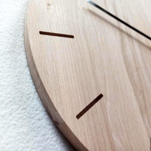 May include: Close-up of a wooden clock face with a light brown finish. The clock has two dark brown rectangular cutouts and black clock hands. The clock is mounted on a white surface.