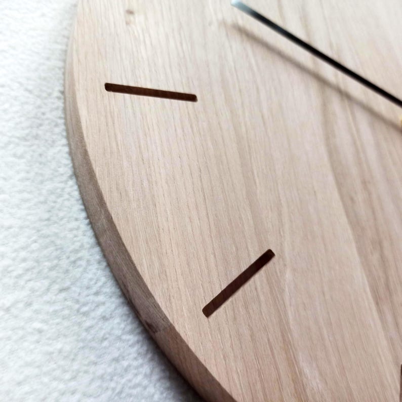 May include: Close-up of a wooden clock face with a light brown finish. The clock has two dark brown rectangular cutouts and black clock hands. The clock is mounted on a white surface.