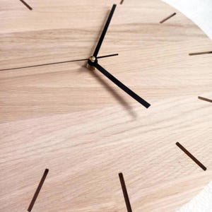 May include: A close-up of a wooden clock face with black hands and hour markers. The clock has a light wood grain finish. The hands are positioned to show the time. The clock is a decorative item.