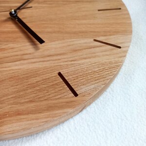 以下が含まれることがあります： 明るい茶色の仕上げの丸い木製時計。文字盤には、シンプルな切り抜きの時間マーカーと黒い針があります。時計は木製で、自然な木目模様があります。